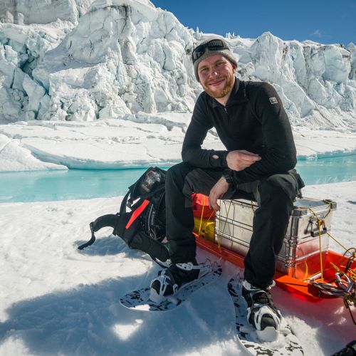 Aapo Rainio kuvattuna Grönlannissa, hän istuu pulkassa ja jäävuori näkyy takana.
