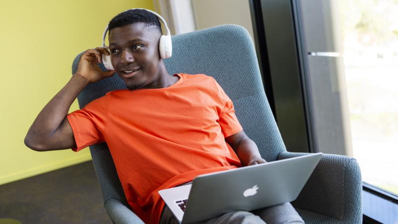 Opiskelija istuu nojatuolissa kannettava tietokone sylissään ja kuulokkeet päässään.