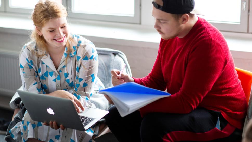 Kaksi henkilöä katsoo tietokoneen näyttöä ja lehtiötä