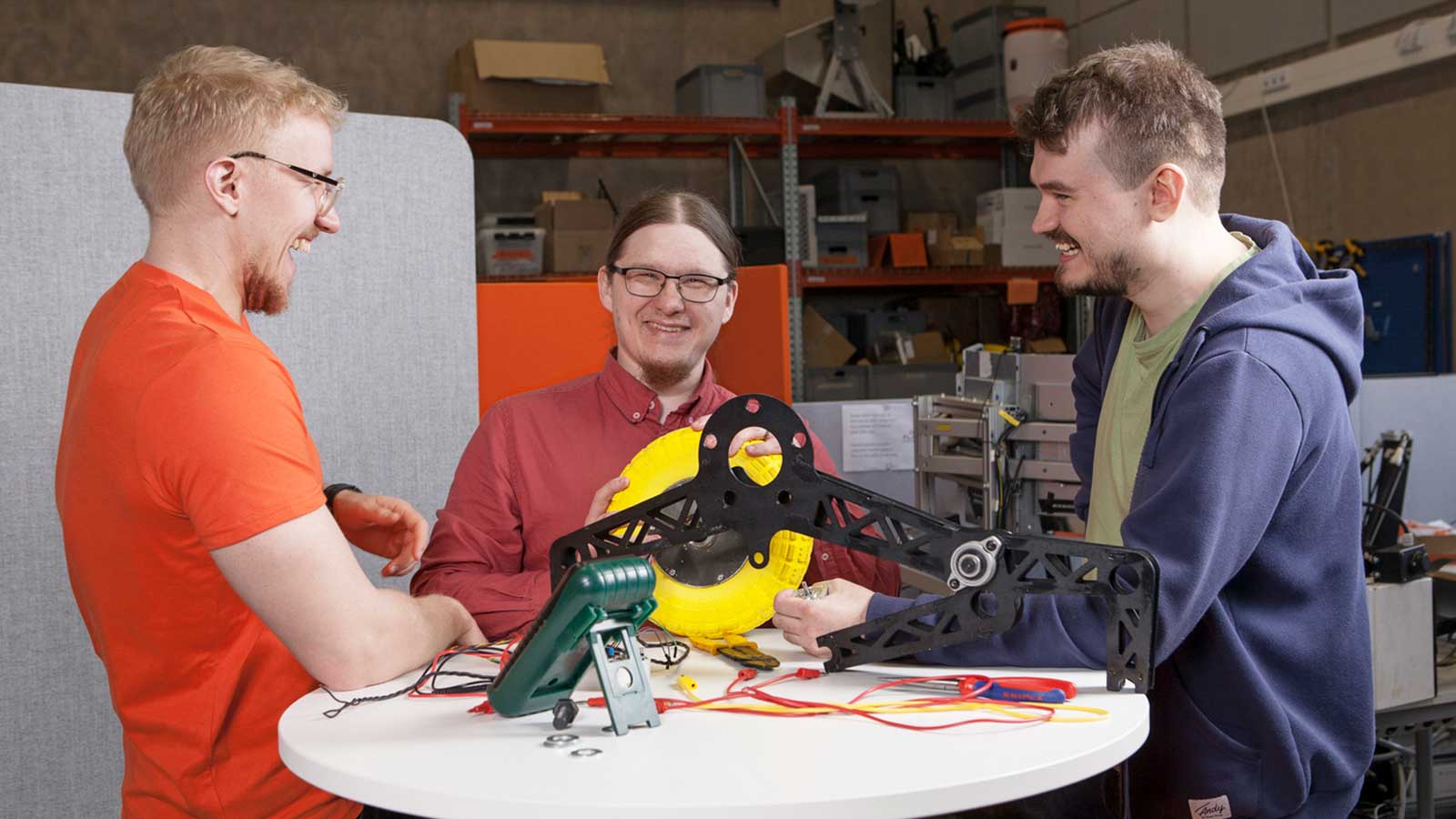 Kolme opiskelijaa rakentaa robottia pöydän ääressä.