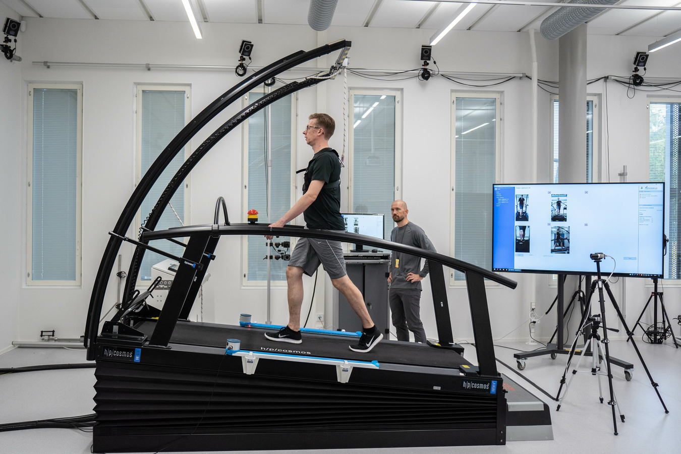 Man walking on treadmil, behind the treadmill two movement analysis cameras and big screen. An other man standing and watching the situation.