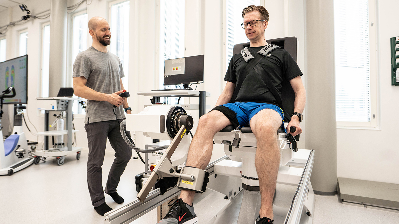 Man sits in isokinetic dynamometer, right lower limb fixed by strap to dynamometer. An other man stands holding emergency switch.