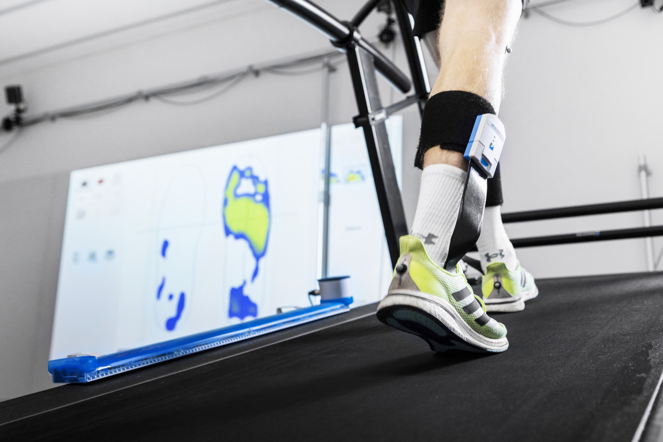 Man walking wearing (picture includes only feet) sport shoes, plantar pressure insoles inside shoes. Colourful plantar pressure figure can be seen on the wall behind the feet.