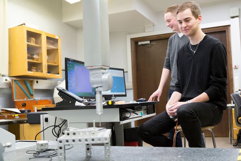 Kaksi konetekniikan opiskelijaa seuraa automatisoidun koneen työskentelyä luokkatilassa. Taustalla tietokoneen näytöllä on ohjelma, jolla konetta ohjelmoidaan.