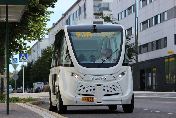 Bussipysäkin katos, robottibussi kadulla, sisällä matkustajia, taustalla Vuosaaren Aurinkolahden katukuvaa.