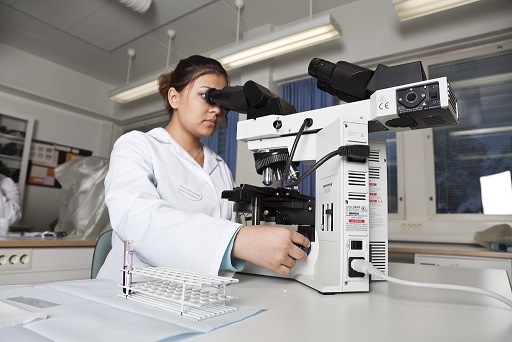 Valkoiseen laboratoriotakkiin pukeutunut nainen käyttää mikroskooppia luokkatilassa.