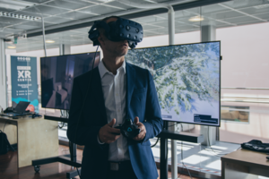 Pukuun pukeutunut mies pitää päässään virtuaalitodellisuuslaseja ja kädessään peliohjainta. Taustalla on iso televisio, joka näyttää kuvaa pelistä, jota mies pelaa virtuaalitodellisuuslasien läpi.