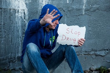 Mies istuu maassa nojaten betoniseinään kädessään lappu, jossa lukee "Stop drugs".
