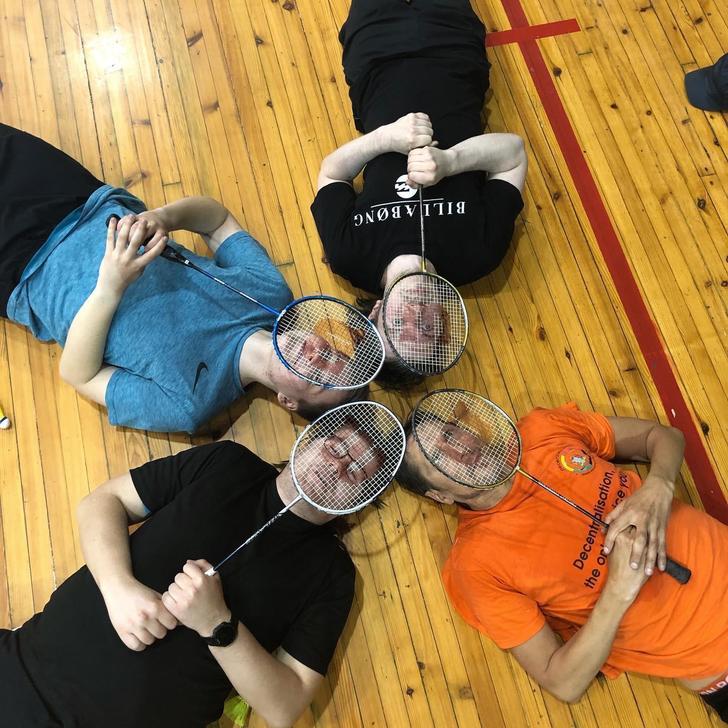 Ylhäältä kuvattuna neljä henkilöä lattiaalla selällään makoulemassa päät vastakkain. He muodostavat tähtikuvion ja heidän kasvonsa edessä on sulkapallomailat.
