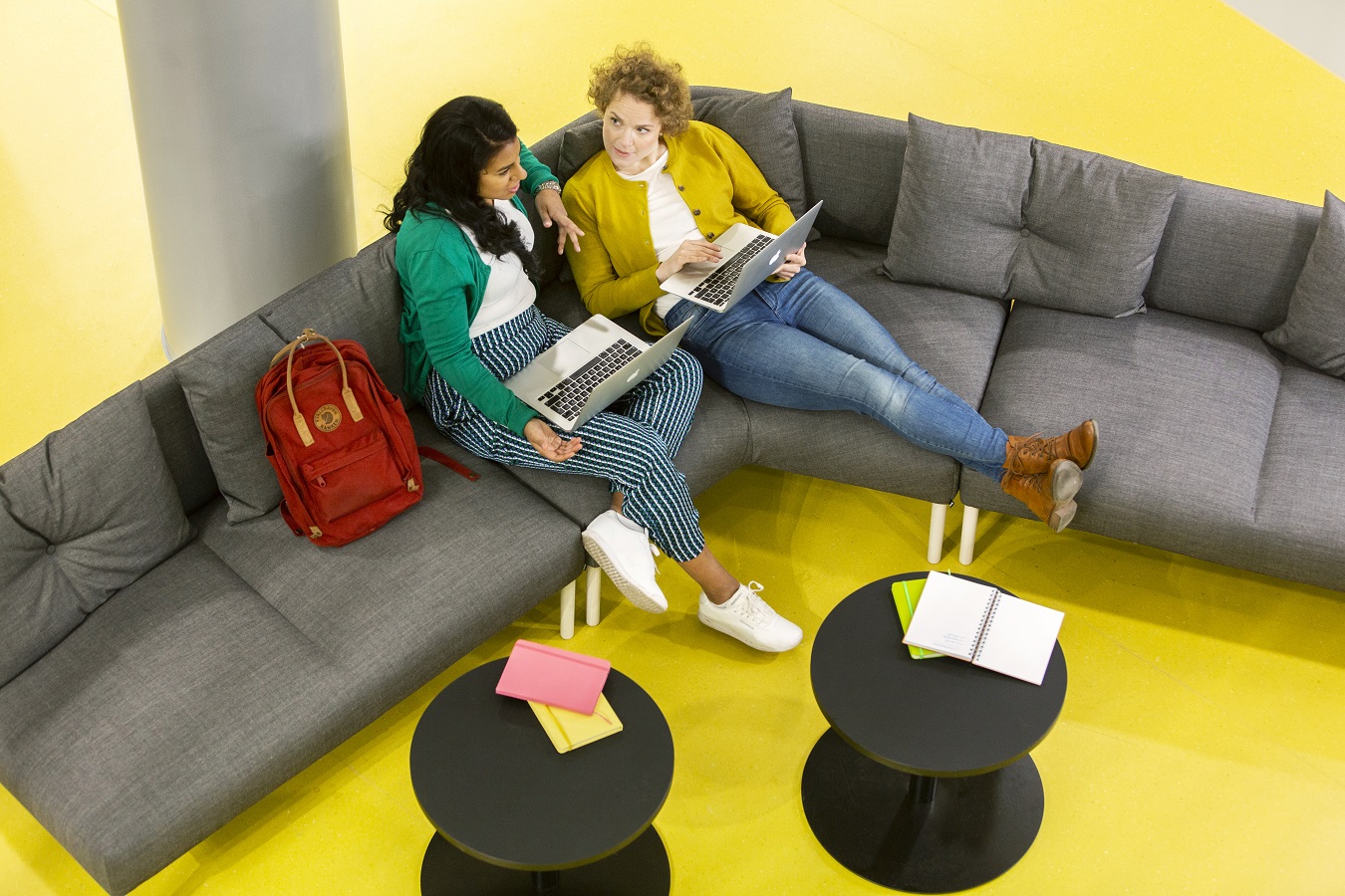 Two people sitting on a sofa with laptops, studying and talking together in a bright lounge area.