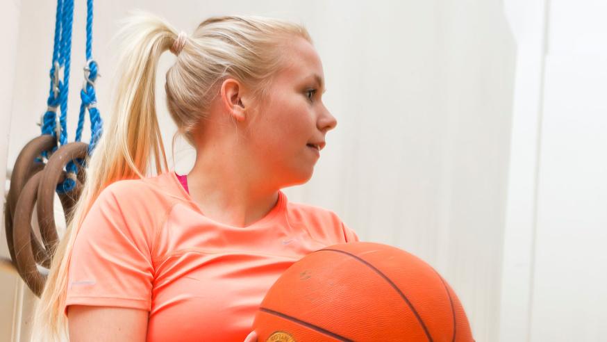Opiskelija pelaa koripalloa liikuntasalissa.