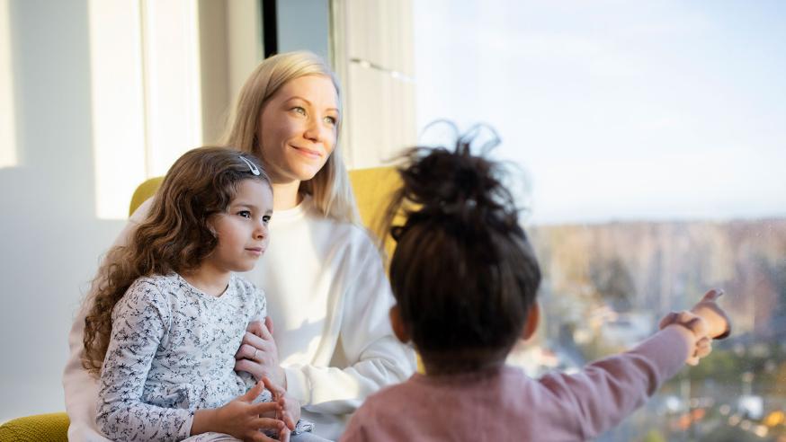 Varhaiskasvatuksen opettaja istuu lapsi sylissään ja toinen edessään ikkunan äärellä.