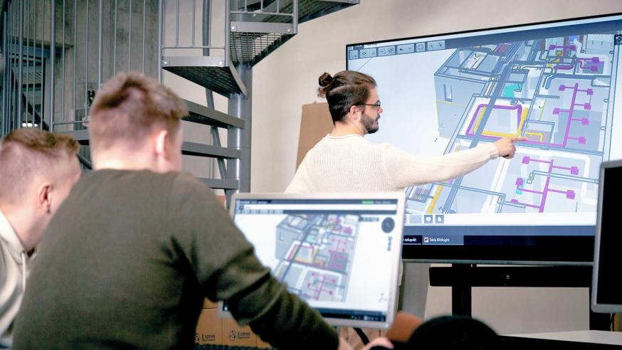 One student showing things on big screen, two students looking at computer screen.