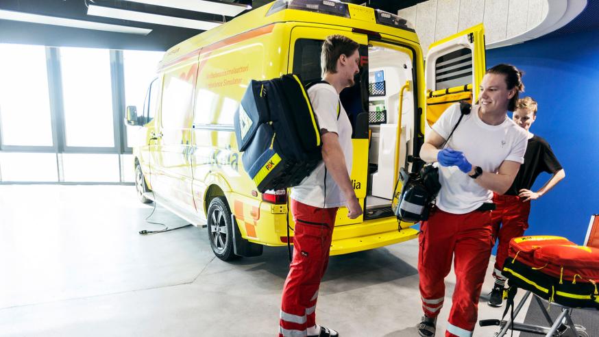 Kolme ensihoidon opiskelijaa ambulanssisimulaattorin edessä Myllypuron kampuksella.