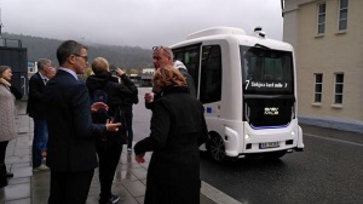 Group of people standing on the street, white robotbus in middle, buildings and grey skies in the background