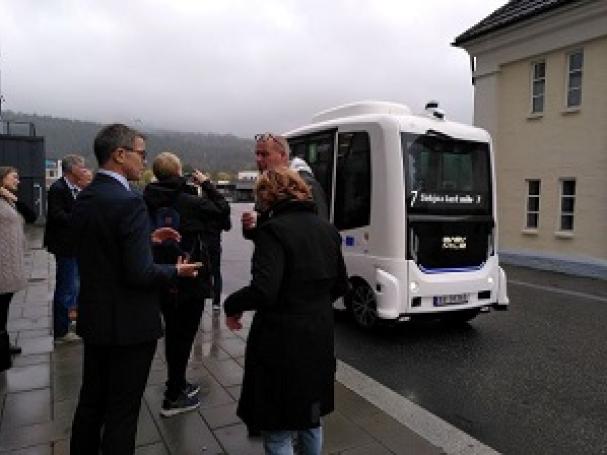 Group of people standing on the street, white robotbus in middle, buildings and grey skies in the background