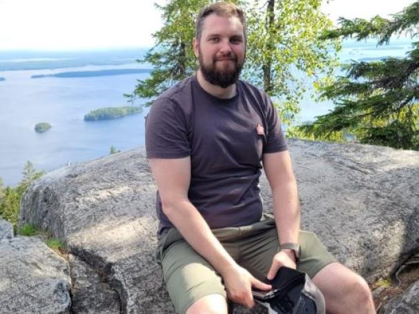 Mika Saastamoinen istumassa kalliolla luonnossa, taustalla näkyy koivu ja järvi.