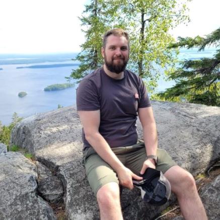 Mika Saastamoinen istumassa kalliolla luonnossa, taustalla näkyy koivu ja järvi.